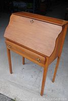 Closed view of vintage drop down desk showing curved front, ash wood finish, brass ring pulls on drawer and drop front.