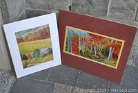 Two matted prints leaning on stone wall: one with red birch trees, the other with barns and fields.