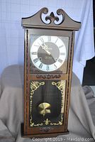 Full frontal view of vintage wooden Citizen wall clock, showing ornate carved top, clock area with Roman numeral face, and glass lower door with gold designs exposing pendulum.