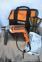 Ridgid drill with side handle, power cord, operation manual, drill bit case, and carrying case displayed together.