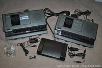 Two gray Sanyo memo-scriber cassette dictation/transcription units side by side with cables, miniature microphones in plastic bag, foot pedal controller, and power adapters on a gray surface.