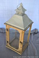 Front angled view of large outdoor decorative lantern showing wood frame, glass panels, and metal roof with decorative top and loop handle.