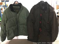 Two men's winter jackets hanging side by side on wooden hangers, one teal green GAP jacket with black collar on left, one dark olive hooded Retreat jacket on right.