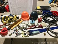 Photo shows a table top with two hard hats (orange and black), yellow and red safety harnesses, various hoses, red tape measure, clear safety glasses, metal clamps, a pressure gauge with hose, and assorted hardware pieces.
