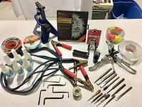 Full overhead view of tools and items including Michelin axle stand, jumper cables, antifreeze bottles, tapes, clamps, drill bits and hex keys laid out on a table.