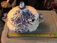 Top and side views of a large ceramic serving dish with blue floral decorations and a scalloped lid, showing scale with a measuring tape, and visible chip damage on lid.