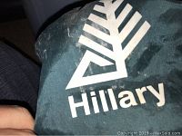 Close-up of green fabric bag with Hillary brand name and logo printed in white.