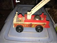 Side angle view of the wooden fire truck toy showing the red and natural wood body, black wheels, firefighter figure in red helmet, silver bell, and mounted ladder.