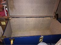 Interior view of the open blue steel trunk showing beige patterned fabric lining and metal latches on sides.