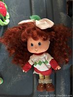 Strawberry Shortcake doll with curly red hair, white bow, red and white dress, yellow striped stockings, and brown shoes.
