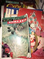 Photo shows a vintage crochet pattern book titled 'Ruffled Doilies' by Coats & Clark, with other crochet and knitting pattern books underneath, including a red cover featuring dolls clothing.