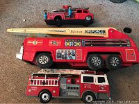 Three vintage fire trucks on carpet including a large red Tonka truck, a medium fire truck with ladder and a smaller tin wind-up truck with fireman figure.