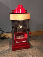Front angled view of the red Hollywood popcorn machine with illuminated marquee and clear glass panels showing interior kettle and accessories.