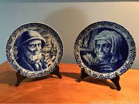 Pair of antique blue Delft plates, old lady and old fisherman, on dark stands against plain background.