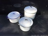 Three white enamelware pots with lids and handles arranged on black surface, showing varying sizes and handle styles