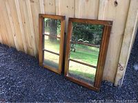 Two antique wood window frame mirrors leaning against a wooden fence on gravel, showing size and overall condition.
