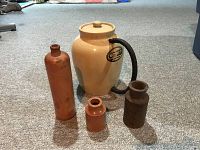 Four antique stoneware pieces on carpet, including a large crock with lid, tall bottle, small jar, and dark crock with spout