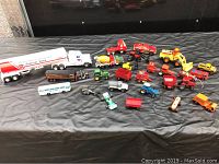 Overview of a collection of over 25 diecast toy tractors, trucks, cars, and heavy equipment in various colors including red, green, yellow, blue, and orange, spread out on a black cloth.