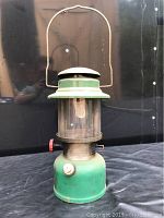 Front view of the vintage green Coleman lantern showing its full height and glass globe, with metal handle raised upright.