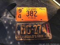Two license plates placed on a black surface showing the Iowa 1964 truck plate with orange background and black text, and the 1964 New York World's Fair black plate with yellow-orange text.