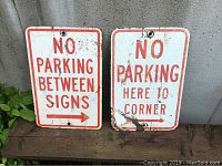 Two metal street signs displayed side by side on a wooden surface against a concrete wall, showing full fronts including text and mounting holes.