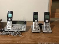 Set of 3 Vtech cordless phones including main base station and three handsets on wooden surface with cords visible.