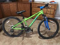 Side profile of the green Raleigh mountain bike with black saddle and blue front suspension fork, showing the entire bike from drivetrain to handlebars and wheels.