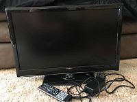 Front view of Axess 24 inch LED TV with remote and power adapter placed in front of the TV on carpet.