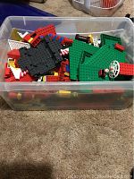 Plastic storage bin filled with assorted LEGO bricks and baseplates in various colors, with some minifigure parts visible.