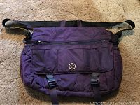 Front view of purple Lululemon bag showing front flap with logo, buckle closures, and adjustable shoulder strap.