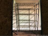 Front view of a chrome 5-shelf wine rack showing wave-shaped holders for horizontal bottle storage, stands on four legs, photographed against a wall with wooden floor.
