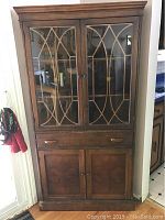 Full front view of antique wood and veneer corner hutch showing double glass doors with decorative muntins, one drawer, and lower wood paneled doors