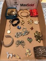 Full view of vintage jewelry lot in box showing wide bangles on top left, black band bracelet, various pins including bow-shaped sterling pin, floral and animal shapes.