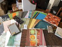 Variety of 12x12 scrapbooking papers spread on a table showing different patterns including florals, geometrics, and stripes; visible alphabet sticker sheet and decorative box.