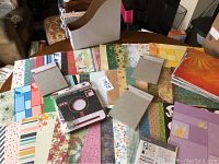 Wide view of scrapbooking papers spread on table showing variety of patterns and colors, plus a scrapbooking accessory kit and embossing folders.