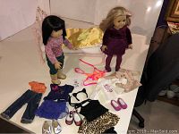 Two American Girl dolls displayed with metal bed and various doll clothes and shoes laid out.