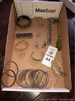 Overall view of jewelry lot showing various bracelets, a feather necklace, earrings, and rings inside a cardboard box with measurement scale.