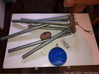 Wind chimes laid flat showing six galvanized metal tubes, oblong wooden striker, round wood top piece, and blue circular hanging decor.