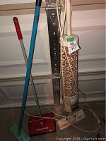 Vintage beige upright vacuum with brown geometric pattern, red and metal manual carpet sweeper, blue mop, and red broom leaning against garage door