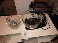 Sunbeam chrome and black stand mixer with power cord on table next to two glass bowls, with one bowl holding two pairs of beaters.
