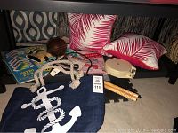 Lot items on floor under table including outdoor pillows, anchor bag, vintage football, sandwich toaster, placemats, coasters, clothesline and jump rope