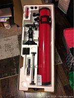 Opened packaging showing the red main telescope tube, tripod mount, and various accessories including eyepieces and adjustment parts arranged in white foam insert.