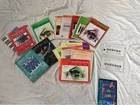 Top-down view of an assortment of music books spread out on a fabric surface. Books include various titles and colors, some with piano-related covers.