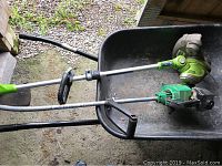 Two grass trimmers placed in a wheelbarrow, Weed Eater gas trimmer on top, Greenworks electric trimmer below, visible wear especially on Weed Eater, missing bottom spool on Weed Eater.