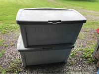 Two gray plastic deck boxes stacked outdoors on a gravel area with grass background, showing weathered lids and small wheels on one side.