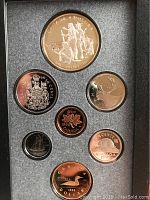 View of seven coins in protective casing including one large silver dollar with Kelsey design and smaller coins arranged around it.