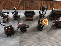 Nine miniature bronze or copper-colored metal collectible pieces arranged on a fabric surface including two souvenir pieces (covered wagon and burro) and seven detailed pencil sharpeners modeled as a telephone, clock, typewriter, lantern, gramophone, candlestick phone, and desk lamp.