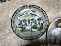 Collector plates displayed on tiled floor, showing a plate with a white porch house, green foliage and floral border.