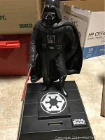 Front view of Darth Vader figure on black base with Imperial emblem and Star Wars logo, holding a red lightsaber.
