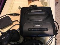 Top view of Sega Genesis black video game console showing cartridge slot, power adapter, official Sega controller, and off-brand controller.